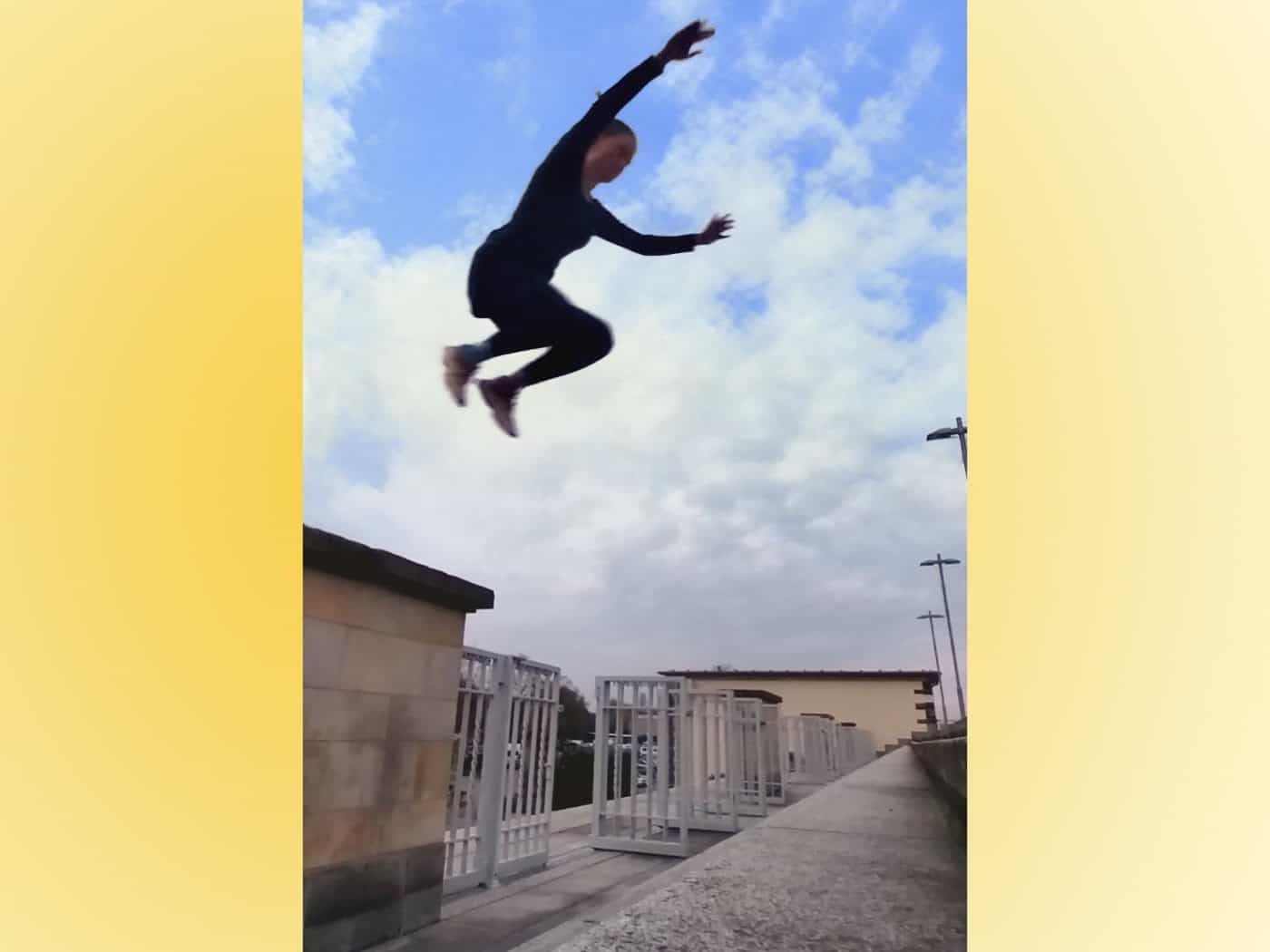 Foto: Person während eines Sprungs in der Luft, vor einem leicht bewölkten Himmel im Hintergrund, unter ihr Treppenstufen