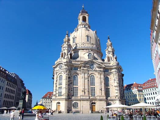 Die Dresdner Frauenkirche in der Totalen vom Neumarkt aus gesehen unter strahlend blauem Himmel. Vor der Kirche steht das Luther-Denkmal; im Vordergrund sind Cafés mit Sonnenschirmen und Passanten zu erkennen. Das Bild zeigt das markante Steingebäude mit seiner großen Kuppel und der Laterne, auf der