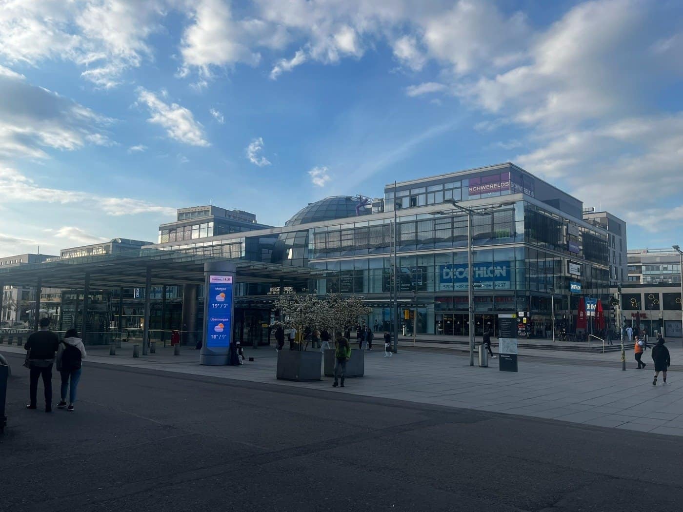 Weitwinkelaufnahme des Wiener Platzes in Dresden bei Tageslicht. Im Hintergrund sind moderne Glasgebäude wie das Kugelhaus und das Gebäude mit dem Decathlon-Store zu sehen. Passanten laufen über den gepflasterten Platz vor dem Hauptbahnhof unter einem blauen, leicht bewölkten Himmel.