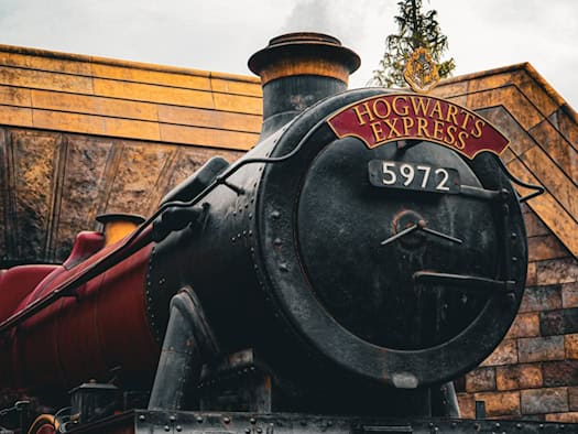 Nahaufnahme der schwarzen Dampflokomotive „Hogwarts Express“ mit der Nummer 5972. Dampf steigt von den Rädern auf, im Hintergrund ist eine rustikale Bahnhofsmauer aus Stein zu sehen. Das Bild steht symbolisch für die Harry Potter Wanderausstellung in Dresden.