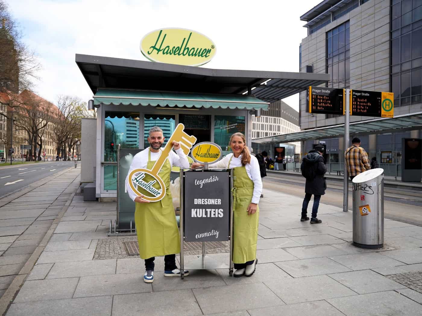 Max Rothe und Annett Walter in grünen Schürzen vor dem Haselbauer-Eispavillon am Dr.-Külz-Ring in Dresden. Rothe hält einen goldenen Schlüssel mit Haselbauer-Logo, daneben ein Aufsteller mit der Aufschrift „Dresdner Kulteis".