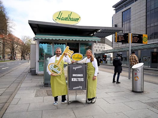Max Rothe und Annett Walter in grünen Schürzen vor dem Haselbauer-Eispavillon am Dr.-Külz-Ring in Dresden. Rothe hält einen goldenen Schlüssel mit Haselbauer-Logo, daneben ein Aufsteller mit der Aufschrift „Dresdner Kulteis".