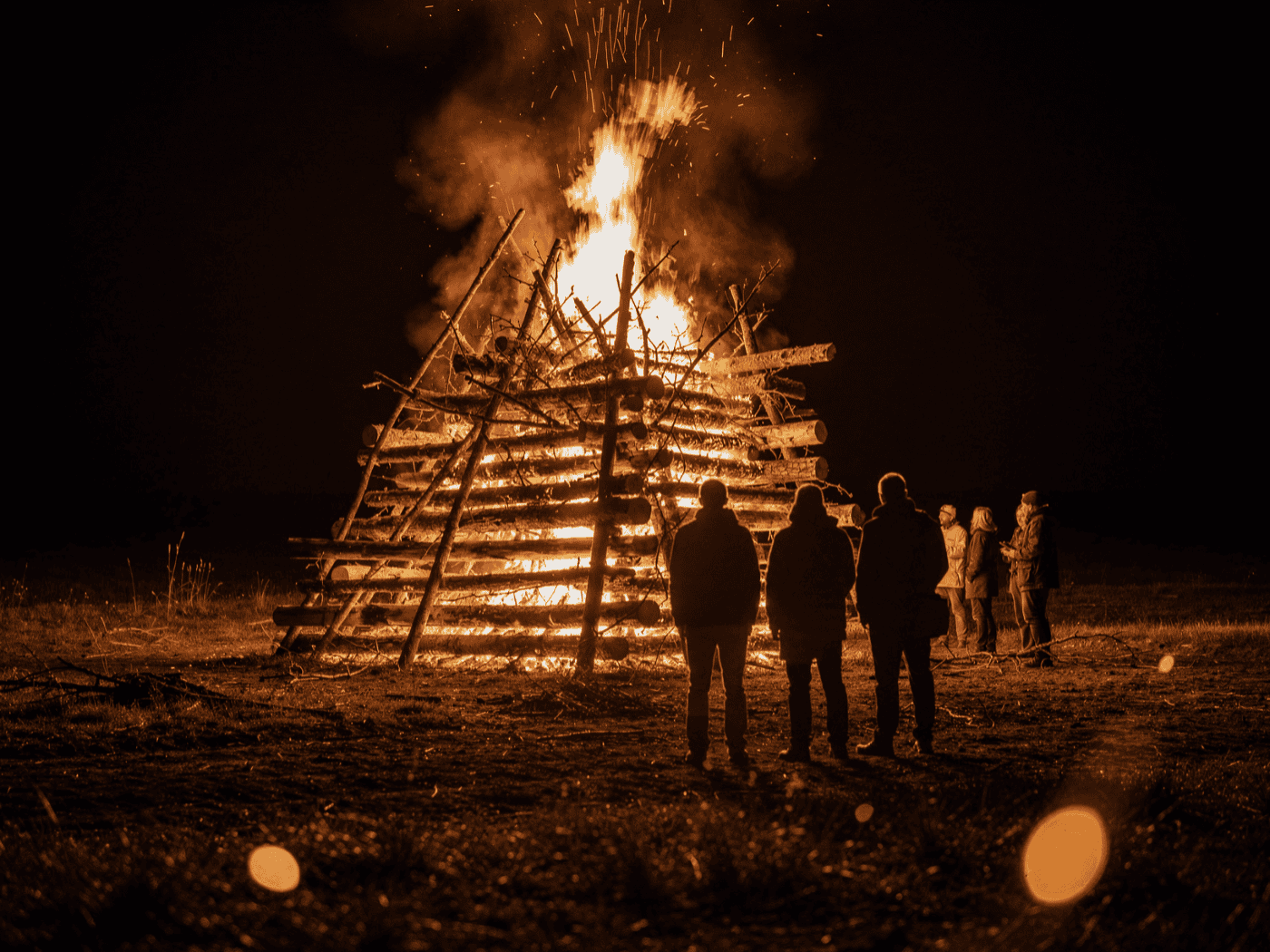 Großes Lagerfeuer bei Nacht, hell lodernde Flammen eines brennenden Holzstapels beim Hexenbrennen zur Walpurgisnacht, im Vordergrund unscharf mehrere Personen als Silhouetten gegen das Feuer.