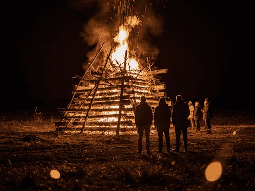 Großes Lagerfeuer bei Nacht, hell lodernde Flammen eines brennenden Holzstapels beim Hexenbrennen zur Walpurgisnacht, im Vordergrund unscharf mehrere Personen als Silhouetten gegen das Feuer.