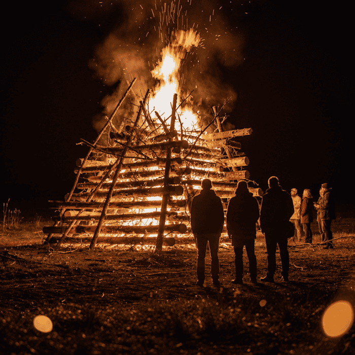 Großes Lagerfeuer bei Nacht, hell lodernde Flammen eines brennenden Holzstapels beim Hexenbrennen zur Walpurgisnacht, im Vordergrund unscharf mehrere Personen als Silhouetten gegen das Feuer.