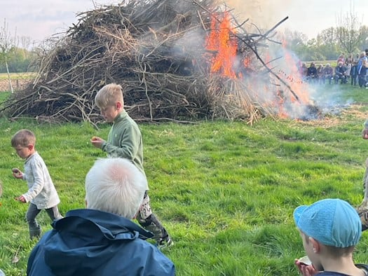 Mehrere Generationen einer Familie sitzen auf einer Holzbank im Grünen und blicken auf ein großes, loderndes Hexenfeuer auf einer Wiese in Dresden. Im Hintergrund sieht man Kinder spielen und weitere Besucher des Volksfestes unter blauem Frühlingshimmel.