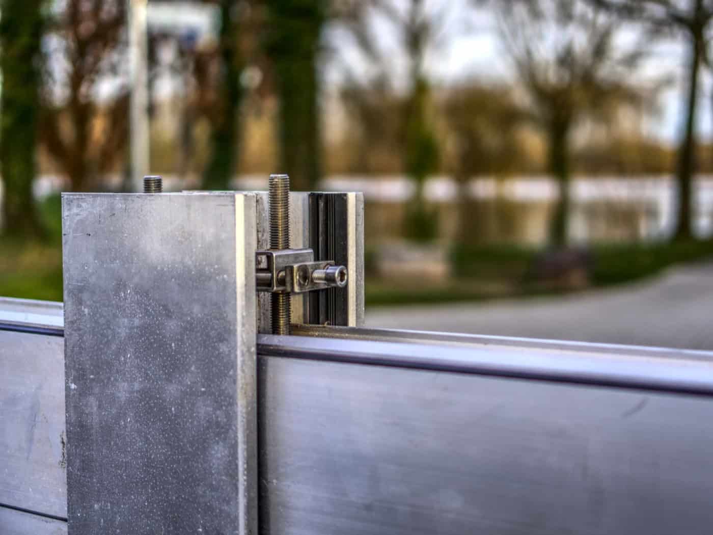 Symbolbild einer mobilen Hochwasserschutzwand aus Metall mit Fokus auf eine mechanische Verschraubung und Halterung. Im Hintergrund ist eine unscharfe Naturkulisse an einem Gewässer zu sehen.