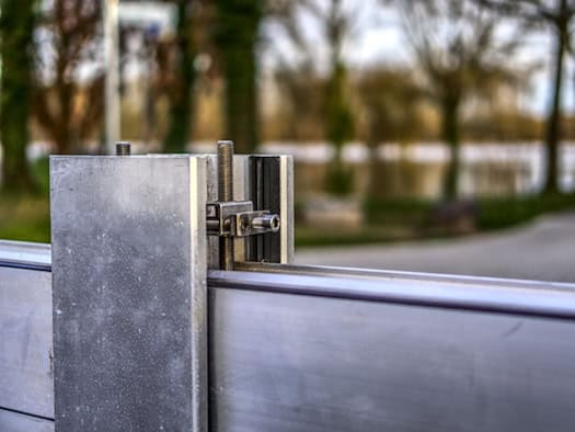 Symbolbild einer mobilen Hochwasserschutzwand aus Metall mit Fokus auf eine mechanische Verschraubung und Halterung. Im Hintergrund ist eine unscharfe Naturkulisse an einem Gewässer zu sehen.
