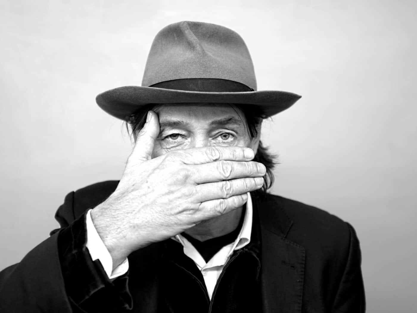 Schwarz-Weiß-Porträt des Künstlers Holger John mit Hut. Er hält eine Hand flach vor seinen Mund, während sein Blick direkt in die Kamera gerichtet ist.