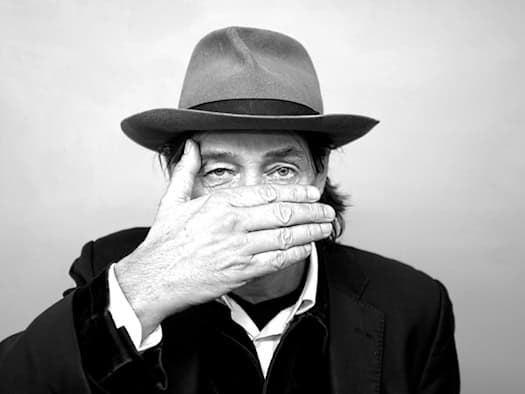 Schwarz-Weiß-Porträt des Künstlers Holger John mit Hut. Er hält eine Hand flach vor seinen Mund, während sein Blick direkt in die Kamera gerichtet ist.