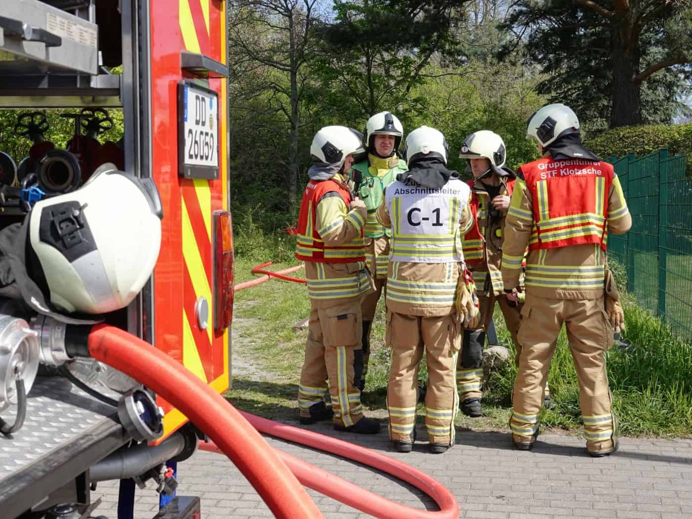 Fünf Feuerwehrleute in sandfarbener Einsatzkleidung und Helmen stehen in einer Gruppe zusammen und besprechen den Einsatz. Einer trägt eine Weste mit der Aufschrift „Abschnittsleiter C-1“, ein anderer die Kennzeichnung „Gruppenführer STF Klotzsche“. Im Vordergrund ist das Heck eines roten Löschfahrz