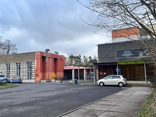 Außenansicht des Förderzentrums „Prof. Dr. Rainer Fetscher“ in Dresden an der Fischhausstraße. Das Bild zeigt den funktionalen Gebäudekomplex mit roter und grauer Fassade, einen Parkplatz im Vordergrund sowie das offizielle Schild der Bildungseinrichtung für körperliche und motorische Entwicklung un