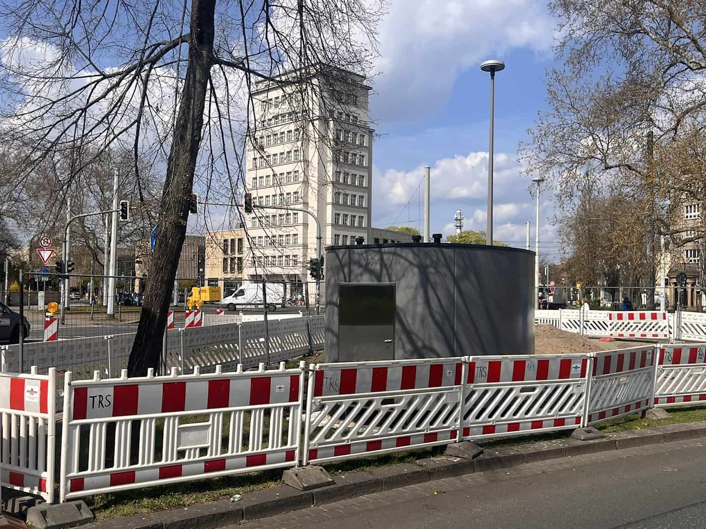 Graue, zylinderförmige öffentliche Toilettenanlage hinter rot-weißen Absperrgittern in Dresden, im Hintergrund ist das markante Hochhaus am Albertplatz zu sehen.