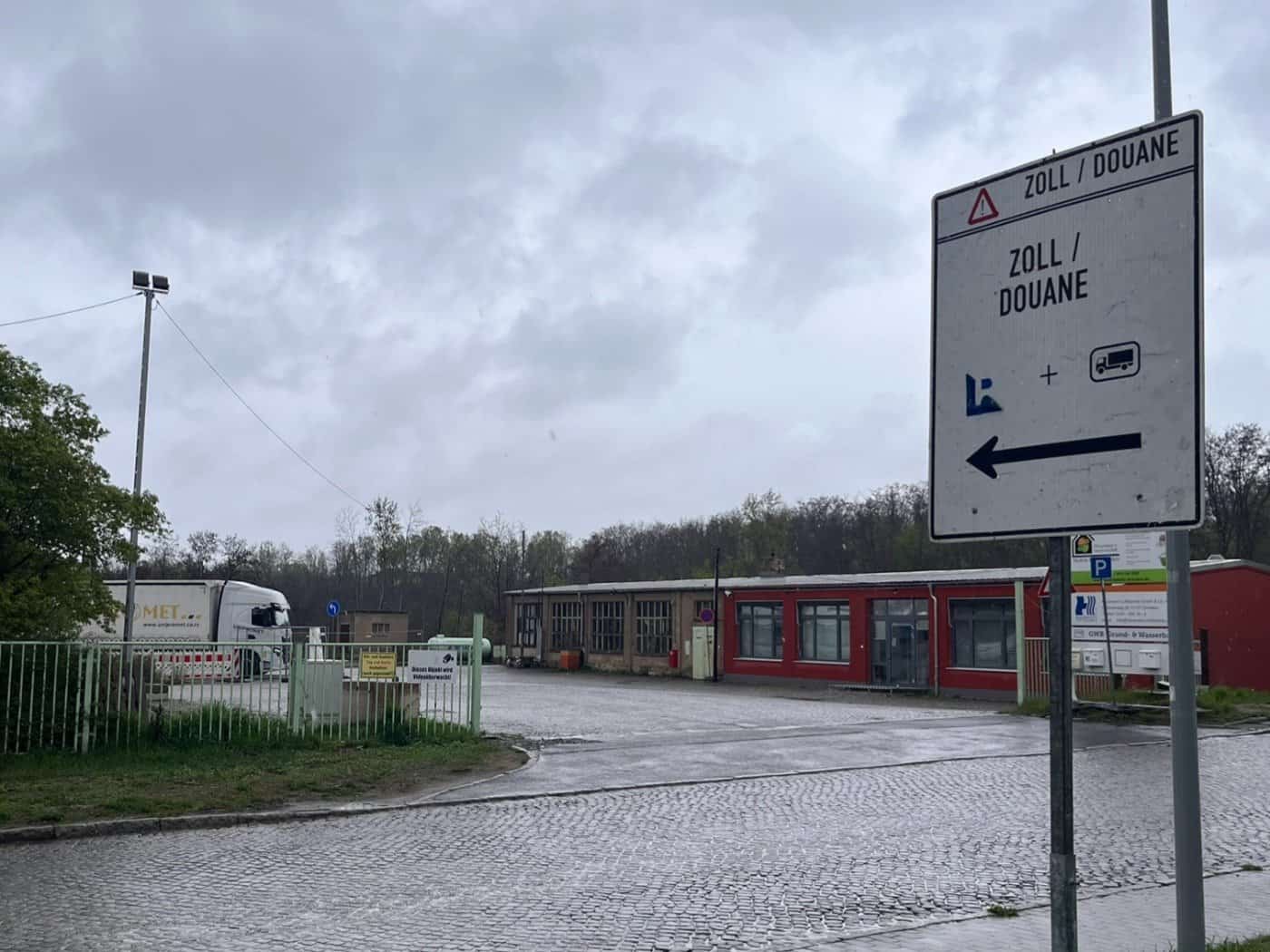 Ein weißes Hinweisschild mit der Aufschrift „ZOLL / DOUANE“ und einem Pfeil nach links steht an einer regnerischen Straße vor einem Gewerbegelände mit Lkw.