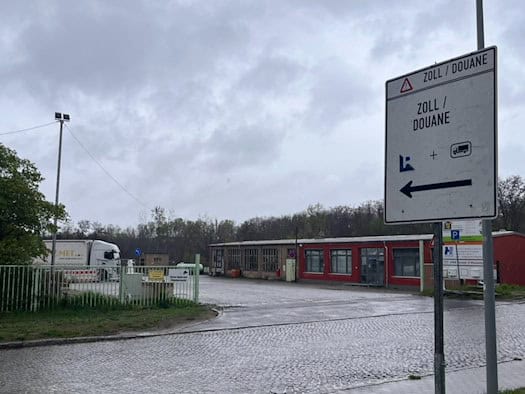 Ein weißes Hinweisschild mit der Aufschrift „ZOLL / DOUANE“ und einem Pfeil nach links steht an einer regnerischen Straße vor einem Gewerbegelände mit Lkw.