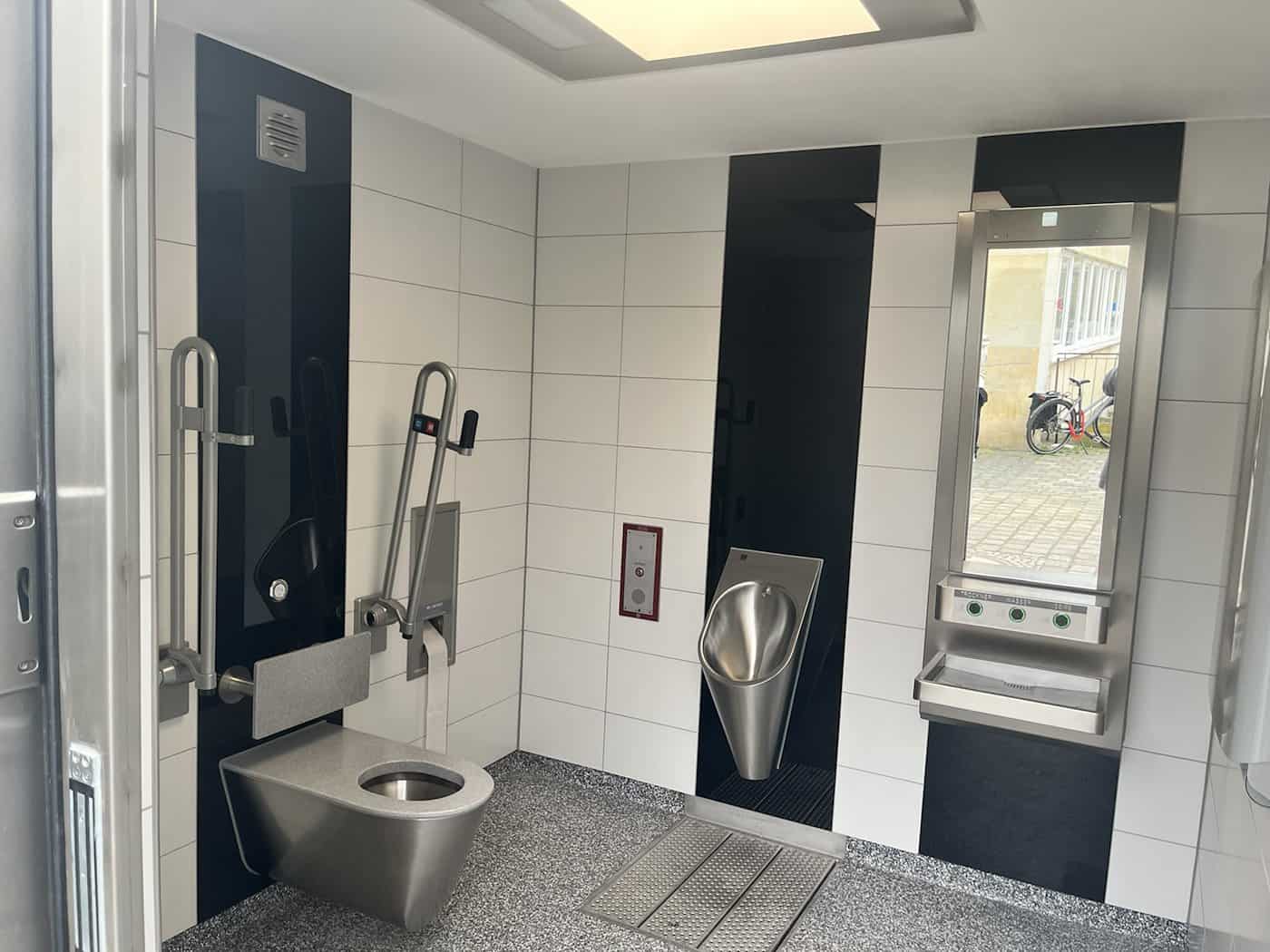 Innenansicht einer modernen barrierefreien öffentlichen Toilette in Dresden mit Edelstahl-WC, Urinal, Haltegriffen und automatischem Waschbecken; helle Fliesen und dunkle Kontrastelemente.