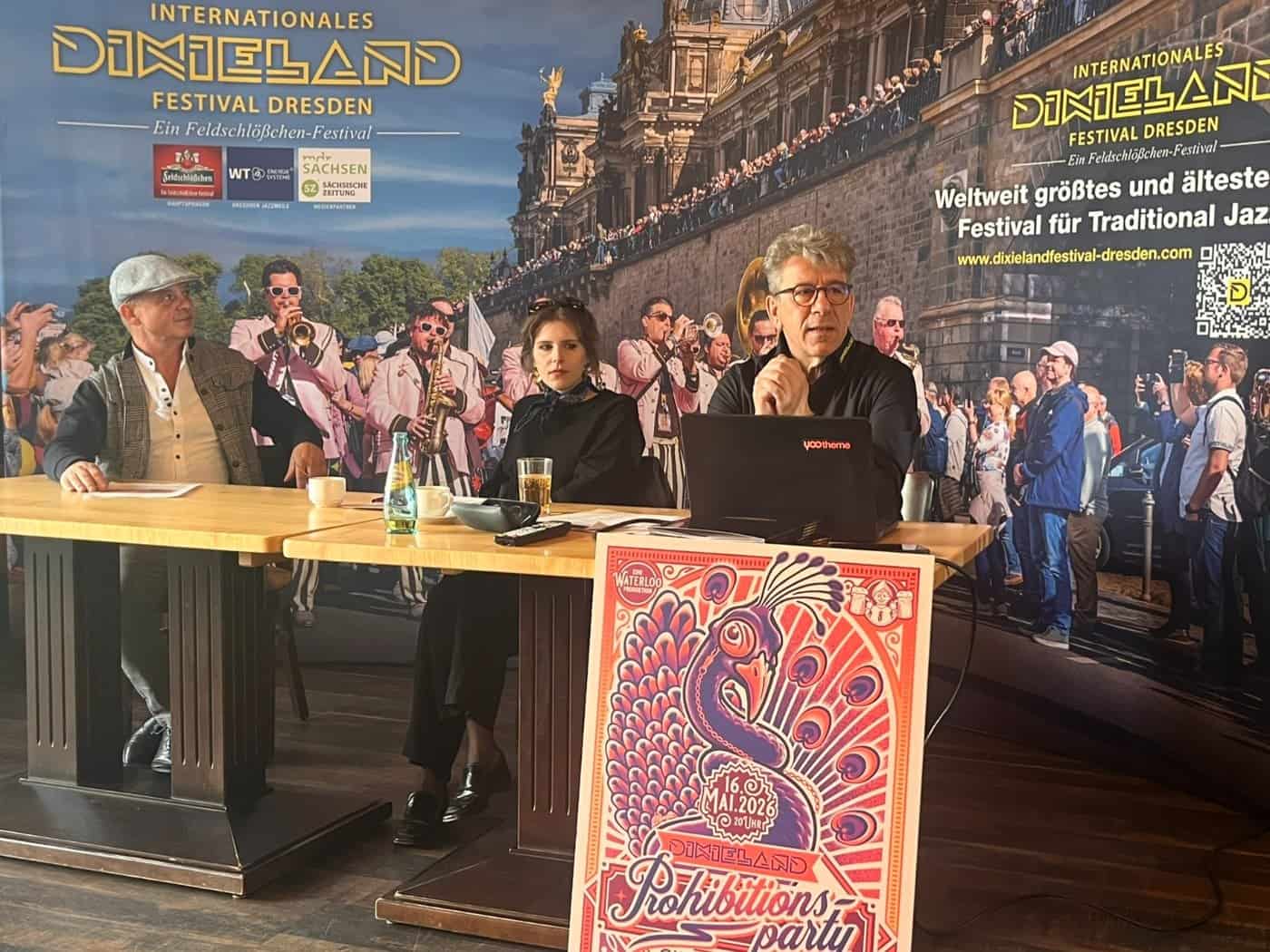 Pressekonferenz zum Internationalen Dixieland Festival Dresden 2026: Drei Organisatoren sitzen an einem Tisch vor einer großen Sponsorenwand; im Vordergrund lehnt ein buntes, illustriertes Plakat für die „Prohibitionsparty – Circus of Swing“.