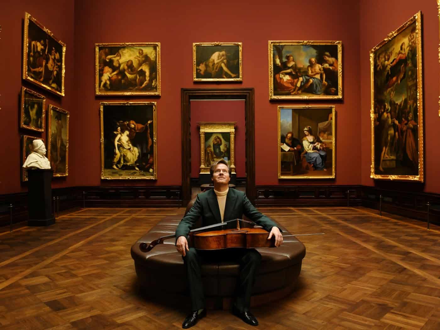 Jan Vogler sitzt mit seinem Cello auf dem Schoß in der Dresdner Gemäldegalerie Alte Meister vor großformatigen historischen Gemälden.