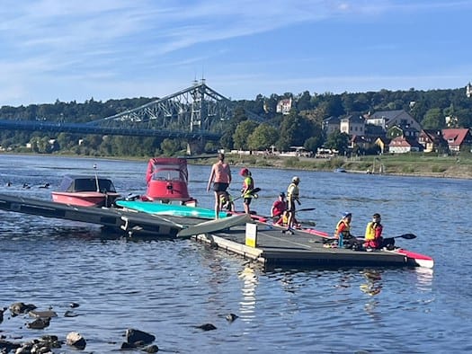 Ein Anlegesteg des Kanu Clubs Dresden an der Elbe. Mehrere Personen in Schwimmwesten bereiten sich auf das Paddeln vor. Im Hintergrund ist die Loschwitzer Brücke (Blaues Wunder) und die bewaldeten Elbhänge mit Häusern zu sehen.