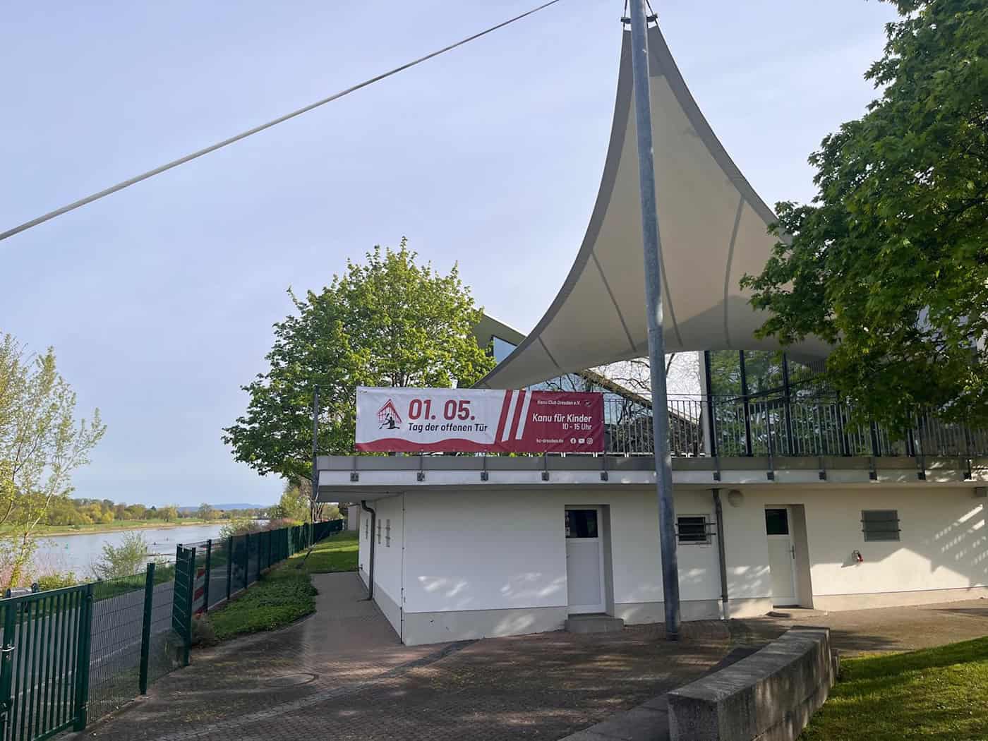 Vereinsgelände des Kanu Club Dresden mit einem Werbebanner für den Tag der offenen Tür am 1. Mai unter einem großen Sonnensegel, im Hintergrund die Elbe.