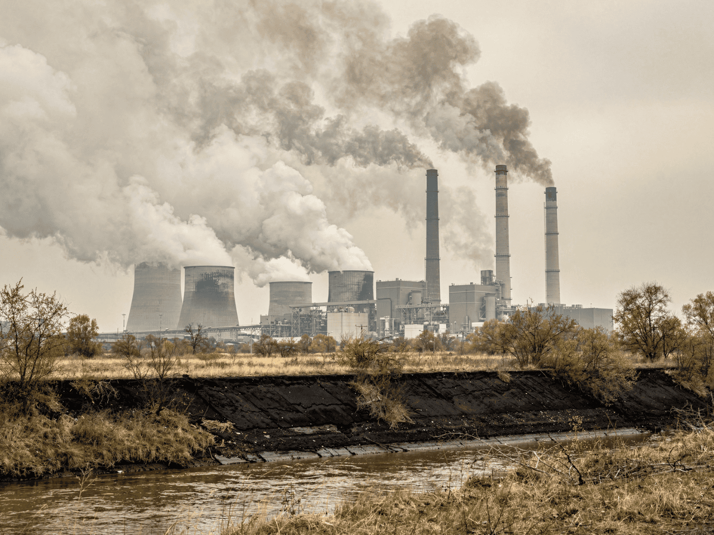 Coal-fired power station with smoking chimneys under a cloudy sky, surrounded by a dry, dusty landscape and murky water in the foreground