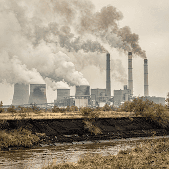 Kohlekraftwerk mit rauchenden Schornsteinen unter bewölktem Himmel, umgeben von trockener, staubiger Landschaft und trübem Gewässer im Vordergrund