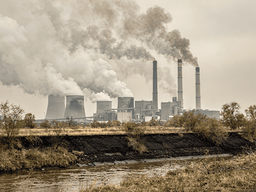 Coal-fired power station with smoking chimneys under a cloudy sky, surrounded by a dry, dusty landscape and murky water in the foreground