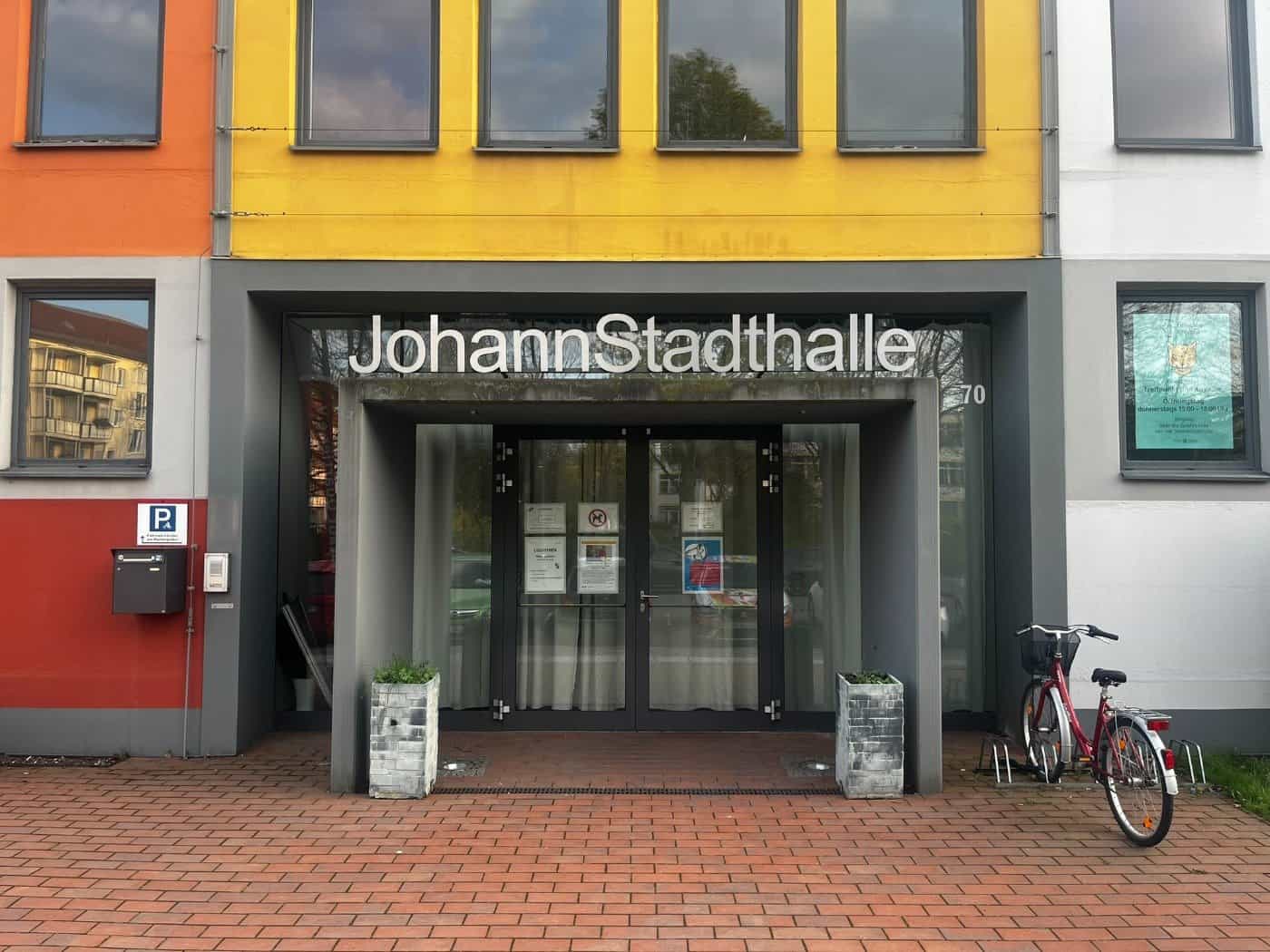 Frontansicht des Eingangs der JohannStadthalle in Dresden. Über der gläsernen Eingangstür prangt der silberne Schriftzug „JohannStadthalle“. Das Gebäude hat eine moderne, farbenfrohe Fassade in Gelb, Orange und Grau. Rechts neben dem Eingang steht ein rotes Fahrrad an einem Fahrradständer auf dem ge