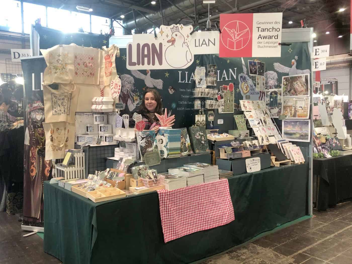 Ein Messestand voller Bücher, Karten und anderer Merchandise-Artikel. Hinter dem Stand steht die Künstlerin LIAN. Oben hängt ein Schild: "Gewinner des Tancho Award der Leipziger Buchmesse".