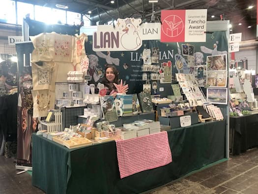 Ein Messestand voller Bücher, Karten und anderer Merchandise-Artikel. Hinter dem Stand steht die Künstlerin LIAN. Oben hängt ein Schild: "Gewinner des Tancho Award der Leipziger Buchmesse".