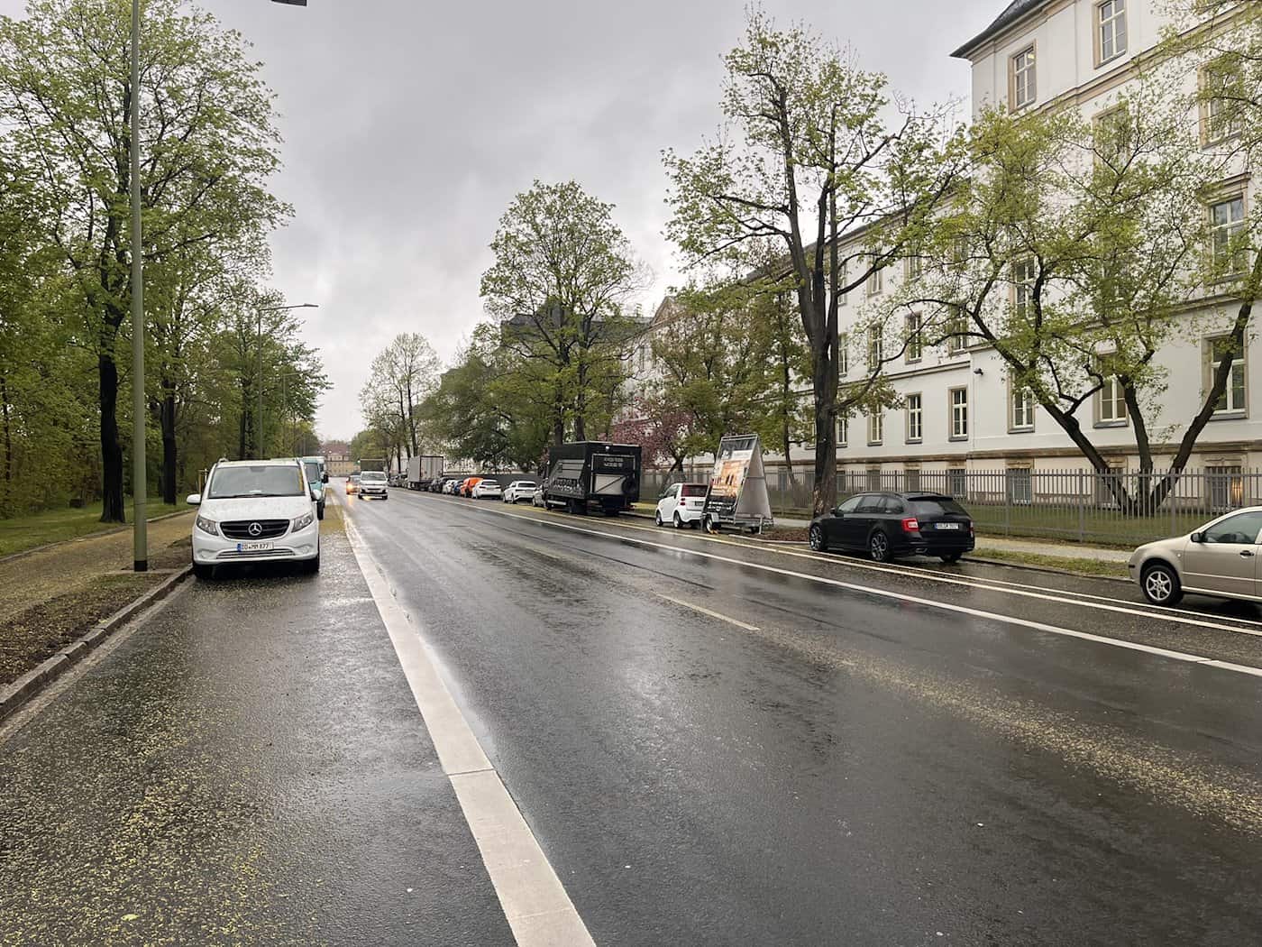 Eine nasse Asphaltstraße (Stauffenbergallee) in Dresden mit am rechten Rand parkenden Pkw und Transportern vor langen, weißen Gebäuden.