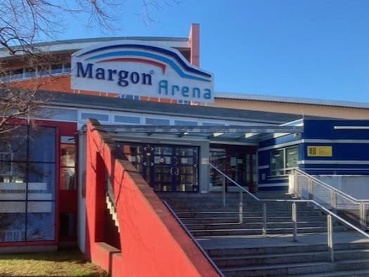 Die Außenansicht der Margon Arena in Dresden mit dem charakteristischen blau-weißen Schriftzug auf dem Dach, einer roten Fassadenseite und der großen Freitreppe zum Eingangsbereich bei blauem Himmel.