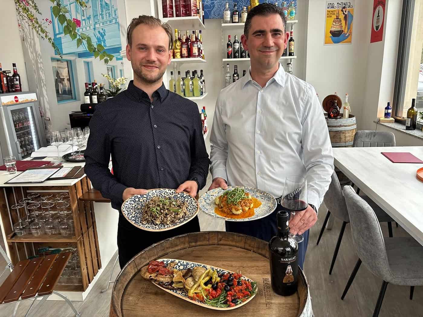 Die Betreiber Sascha Janus und Paul Schlögel stehen mit Tellern voll italienischer Pasta und Antipasti in ihrem hellen Bistro in Dresden. Im Hintergrund sind Regale mit sardischen Weinen und Feinkostprodukten zu sehen.