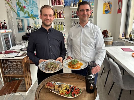 Die Betreiber Sascha Janus und Paul Schlögel stehen mit Tellern voll italienischer Pasta und Antipasti in ihrem hellen Bistro in Dresden. Im Hintergrund sind Regale mit sardischen Weinen und Feinkostprodukten zu sehen.