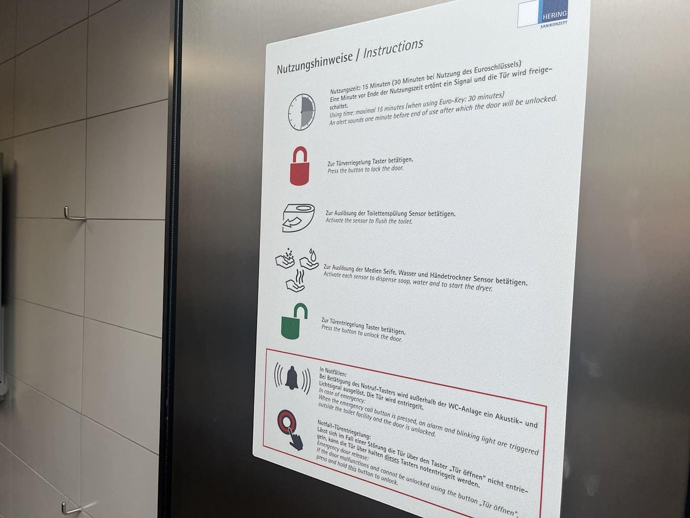 Hinweisschild mit Nutzungshinweisen in einer öffentlichen Toilette in Dresden; Piktogramme für Türverriegelung, Spülung und Notruf auf Deutsch und Englisch.