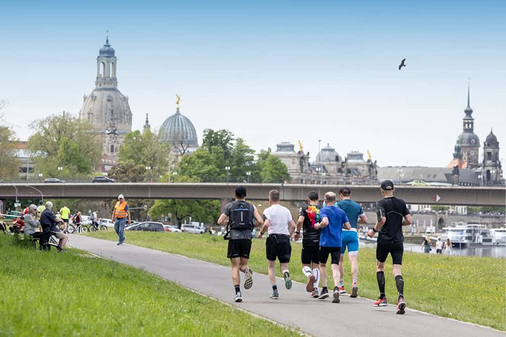Eine Gruppe von Marathonläufern rennt auf dem asphaltierten Elberadweg in Dresden; im Hintergrund ist die berühmte Silhouette der Altstadt mit der Frauenkirche, der Kunstakademie und dem Hausmannsturm zu sehen.