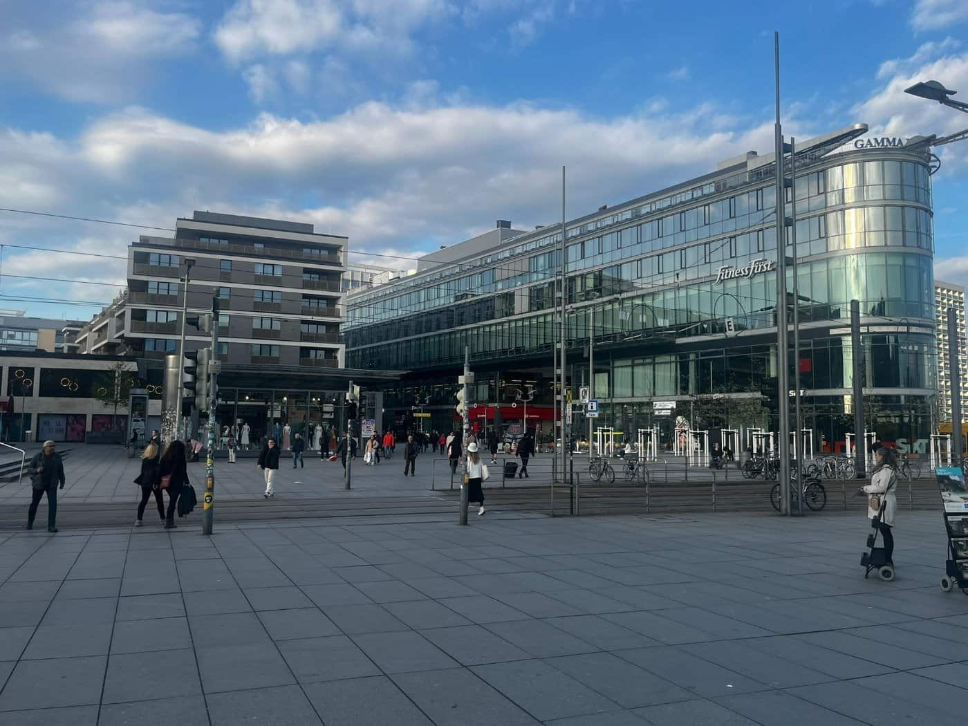 Weitwinkelansicht des Wiener Platzes in Dresden am Übergang zur Prager Straße.  Im Bild sind moderne Geschäftsgebäude mit Glasfassaden (u. a. „Fitness First“ und „GAMMA“) sowie Passanten auf dem weitläufigen, gepflasterten Platz zu sehen.