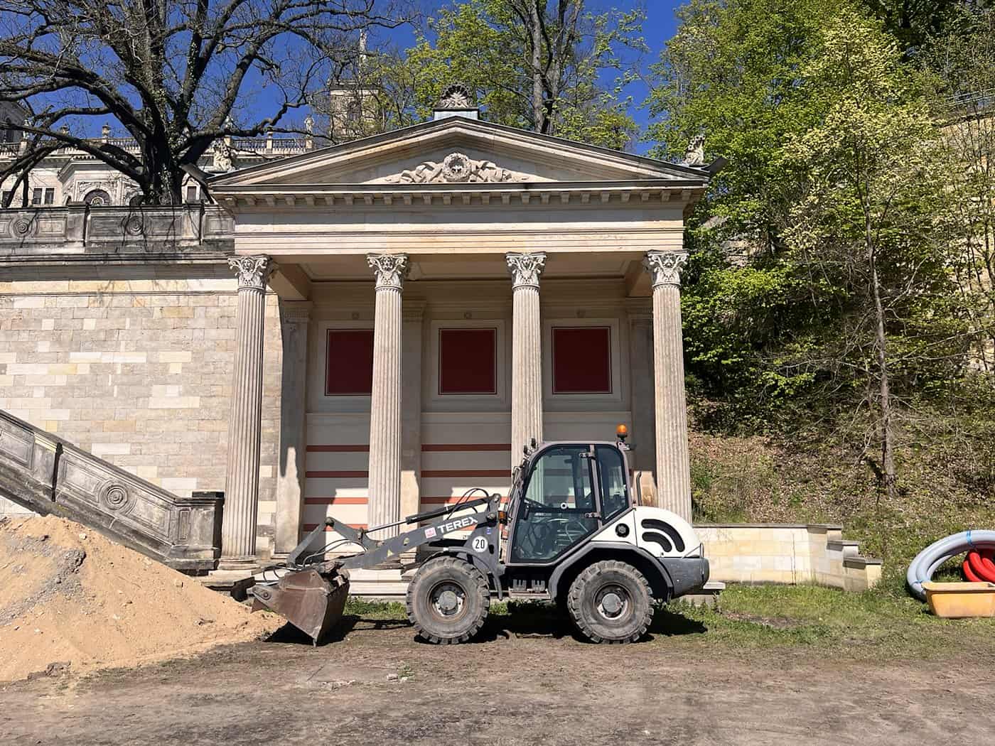 Der bereits sanierte Ostflügel des Römischen Bades in Dresden mit korinthischen Säulen und rötlicher Fassadengestaltung. Im Vordergrund parkt ein kleiner Terex-Radlader neben einem Sandhaufen auf der Baustelle.