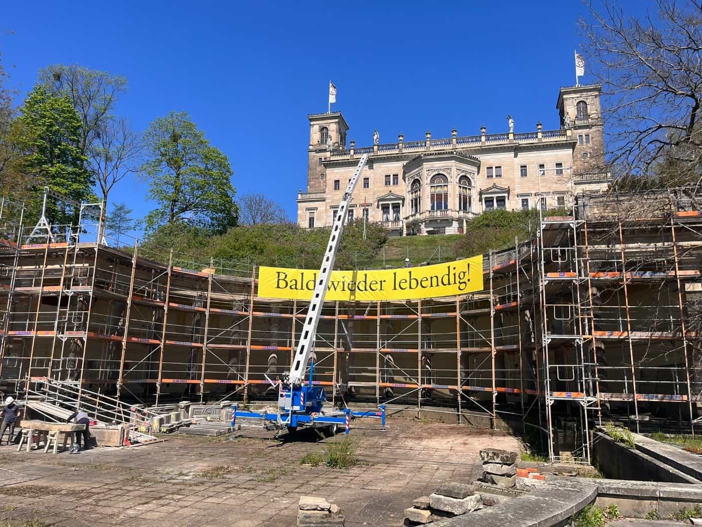 Baustelle am Römischen Bad in Dresden: Ein hohes Baugerüst umschließt die halbkreisförmigen Kolonnaden unterhalb von Schloss Albrechtsberg. Ein gelbes Banner mit der Aufschrift „Bald wieder lebendig!“ hängt am Gerüst, im Vordergrund steht ein blauer Anhängerkran.