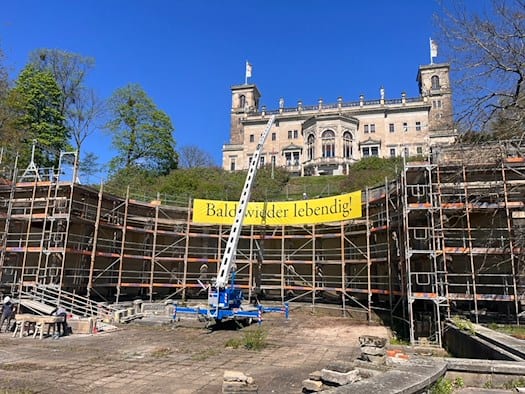Baustelle am Römischen Bad in Dresden: Ein hohes Baugerüst umschließt die halbkreisförmigen Kolonnaden unterhalb von Schloss Albrechtsberg. Ein gelbes Banner mit der Aufschrift „Bald wieder lebendig!“ hängt am Gerüst, im Vordergrund steht ein blauer Anhängerkran.