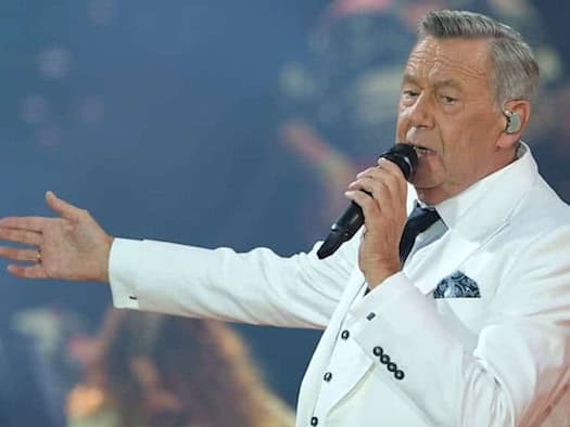 Porträt des Sängers Roland Kaiser während eines Live-Auftritts bei der Kaisermania in Dresden. Er trägt ein weißes Sakko, ein weißes Hemd mit dunkler Krawatte und singt in ein schwarzes Mikrofon vor einer atmosphärisch beleuchteten Konzertkulisse.