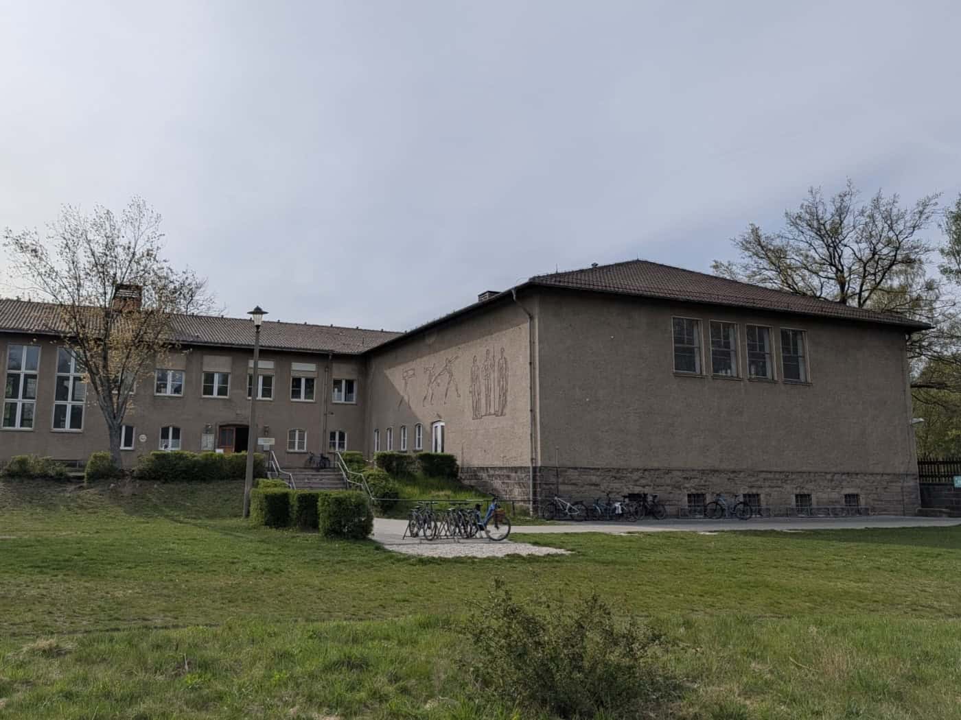 Außenansicht der Schwimmhalle Dresden-Klotzsche an der Königsbrücker Landstraße. Das zweistöckige Gebäude mit grauer Putzfassade und Walmdach zeigt ein Wandbild an der Seitenwand. Vor der Halle befinden sich eine Grünfläche, Fahrradständer und Bäume unter einem leicht bewölkten Himmel.