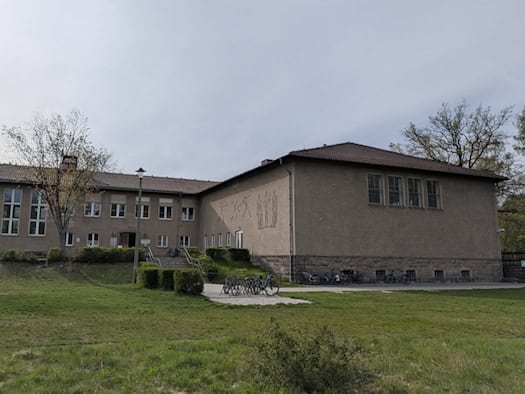 Außenansicht der Schwimmhalle Dresden-Klotzsche an der Königsbrücker Landstraße. Das zweistöckige Gebäude mit grauer Putzfassade und Walmdach zeigt ein Wandbild an der Seitenwand. Vor der Halle befinden sich eine Grünfläche, Fahrradständer und Bäume unter einem leicht bewölkten Himmel.