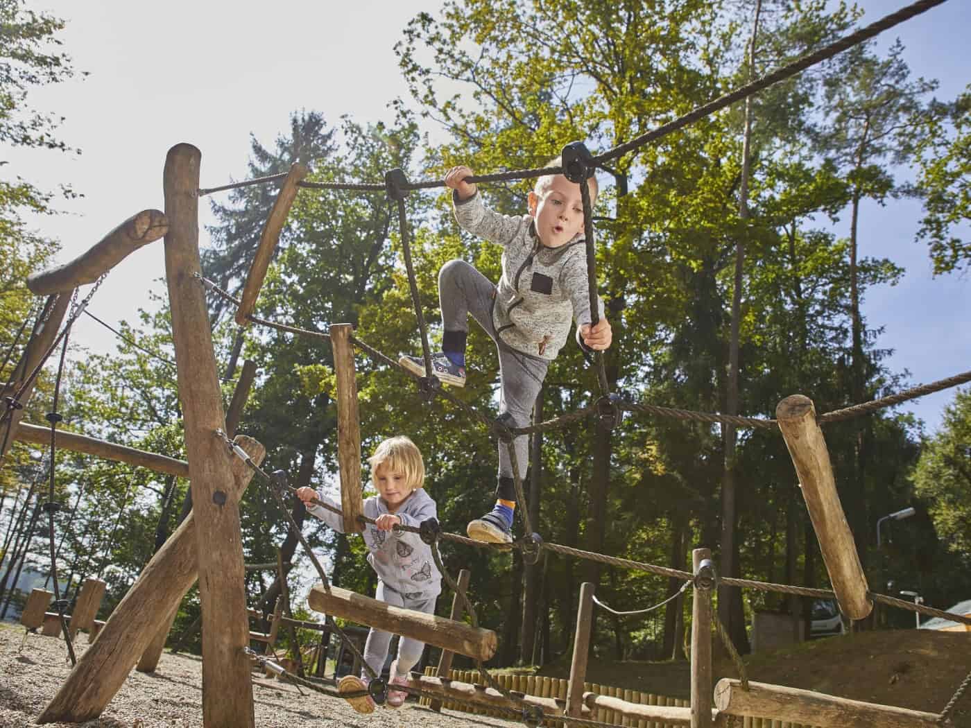 Zwei Kinder klettern auf einem hölzernen Spielgerät mit Kletterseilen in einem bewaldeten Außenbereich. Das Bild dient als Symbolfoto für den geplanten Entdecker-Spielplatz am Frauensteiner Platz.