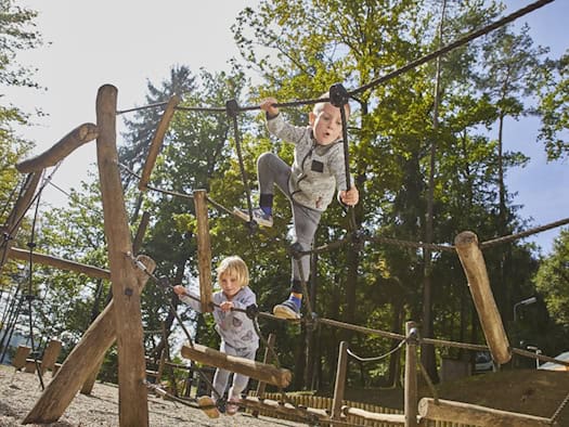 Zwei Kinder klettern auf einem hölzernen Spielgerät mit Kletterseilen in einem bewaldeten Außenbereich. Das Bild dient als Symbolfoto für den geplanten Entdecker-Spielplatz am Frauensteiner Platz.