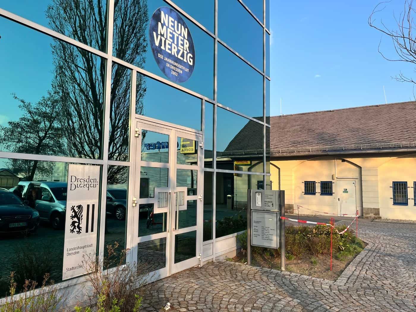 Die moderne Glasfassade des Stadtarchivs Dresden mit dem Logo der Landeshauptstadt und einem runden Aufkleber zur Jahrhundertflut 2002. Rechts im Hintergrund ist ein helles historisches Gebäude mit Ziegeldach zu sehen.