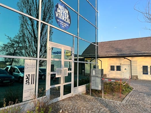 Die moderne Glasfassade des Stadtarchivs Dresden mit dem Logo der Landeshauptstadt und einem runden Aufkleber zur Jahrhundertflut 2002. Rechts im Hintergrund ist ein helles historisches Gebäude mit Ziegeldach zu sehen.