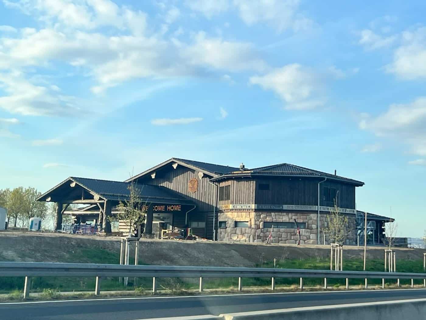 Ein großes, rustikales Blockhaus aus dunklen Baumstämmen mit der Aufschrift „Welcome Home“ steht auf einer Anhöhe neben der Bundesstraße B170 in Bannewitz unter einem leicht bewölkten blauen Himmel.