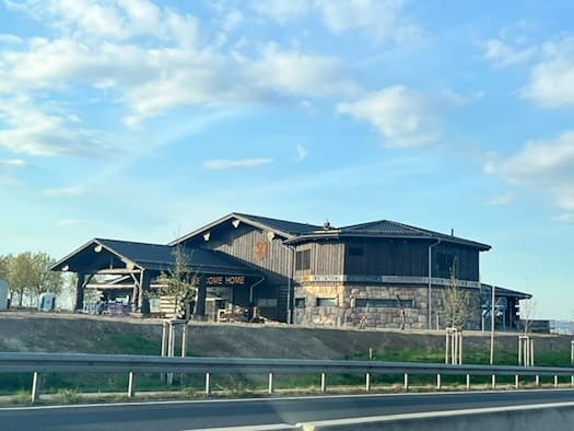 Ein großes, rustikales Blockhaus aus dunklen Baumstämmen mit der Aufschrift „Welcome Home“ steht auf einer Anhöhe neben der Bundesstraße B170 in Bannewitz unter einem leicht bewölkten blauen Himmel.