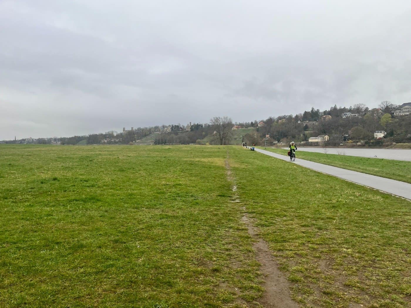 Blick auf den asphaltierten Elberadweg in Dresden bei bewölktem Himmel. Rechts fließt die Elbe, links erstrecken sich die weitläufigen Elbwiesen. Im Hintergrund sind die bewaldeten Elbhänge mit Villen und Schlössern (Albrechtsberg) zu erkennen. Ein einzelner Radfahrer ist auf dem Weg unterwegs.